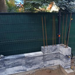 Usługi budowlane - Wznoszenie murku z betonowych bloczków w kolorze szaro-białym, widoczne pręty zbrojeniowe wystające z pustaków, w tle zielona siatka ogrodzeniowa i drzewa.