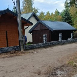 Usługi budowlane - Nowoczesne ogrodzenie z kamiennych bloczków oddzielające posesję z drewnianym garażem i domem od piaszczystej drogi.
