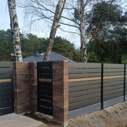 Usługi budowlane - Nowoczesne ogrodzenie z poziomych paneli w kolorze grafitowym z elementami dekoracyjnymi w kolorze drewna, osadzone na słupkach wykończonych imitacją cegły, widok z boku na tle drzew i domu.