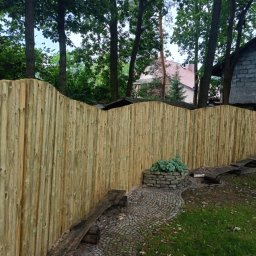 Usługi budowlane - Nowe ogrodzenie z pionowych desek z zaokrąglonym wykończeniem, widok z zewnątrz, z fragmentem ścieżki z kostki brukowej i kamienną rabatką z roślinami.