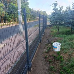 Usługi budowlane - Zamontowane, grafitowe ogrodzenie panelowe przy chodniku z kostki brukowej, z widocznym wiadrem po farbie obok słupka.