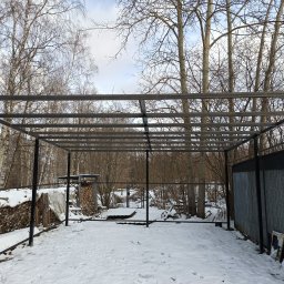 Usługi budowlane - Szkielet metalowej konstrukcji w zimowym krajobrazie, z widocznym stosem drewna i częścią blaszanej ściany w tle.