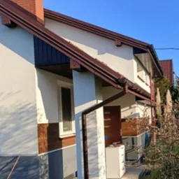 Elewacja domu jednorodzinnego w trakcie odnawiania: widoczna świeżo pomalowana ściana, brązowe rynny, fragment okna, drzwi wejściowe i rozstawione narzędzia malarskie.