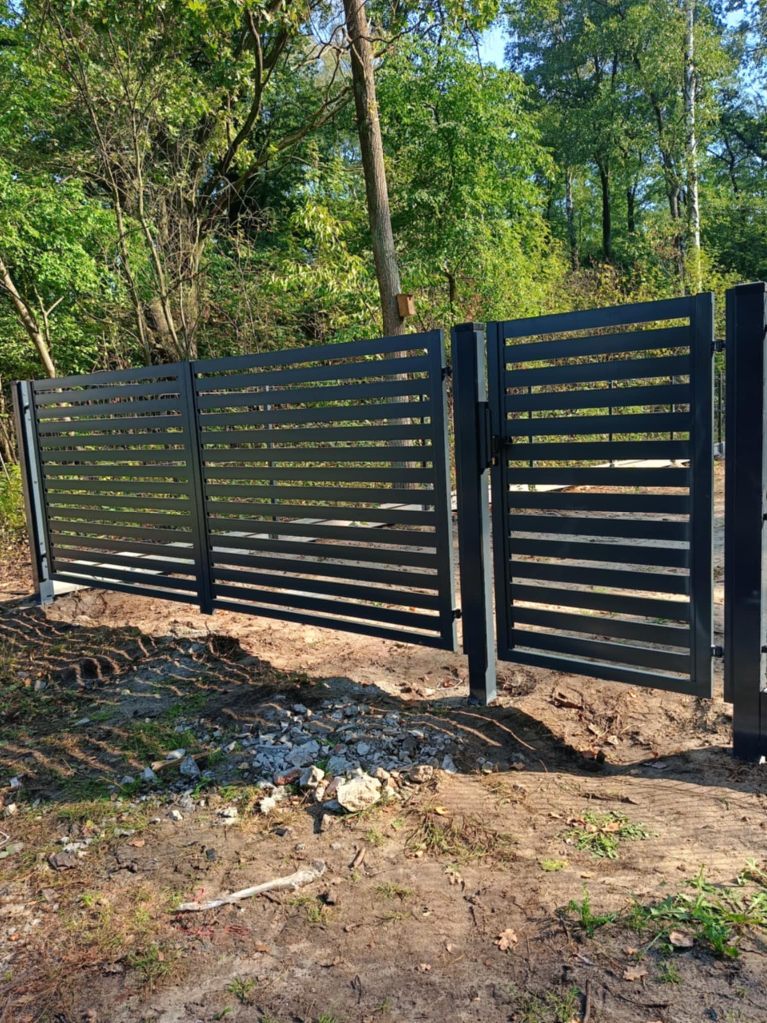 Nowoczesne, poziome, czarne przęsła ogrodzeniowe z furtką, na tle zieleni drzew, świeżo po montażu, widoczne ślady prac ziemnych.