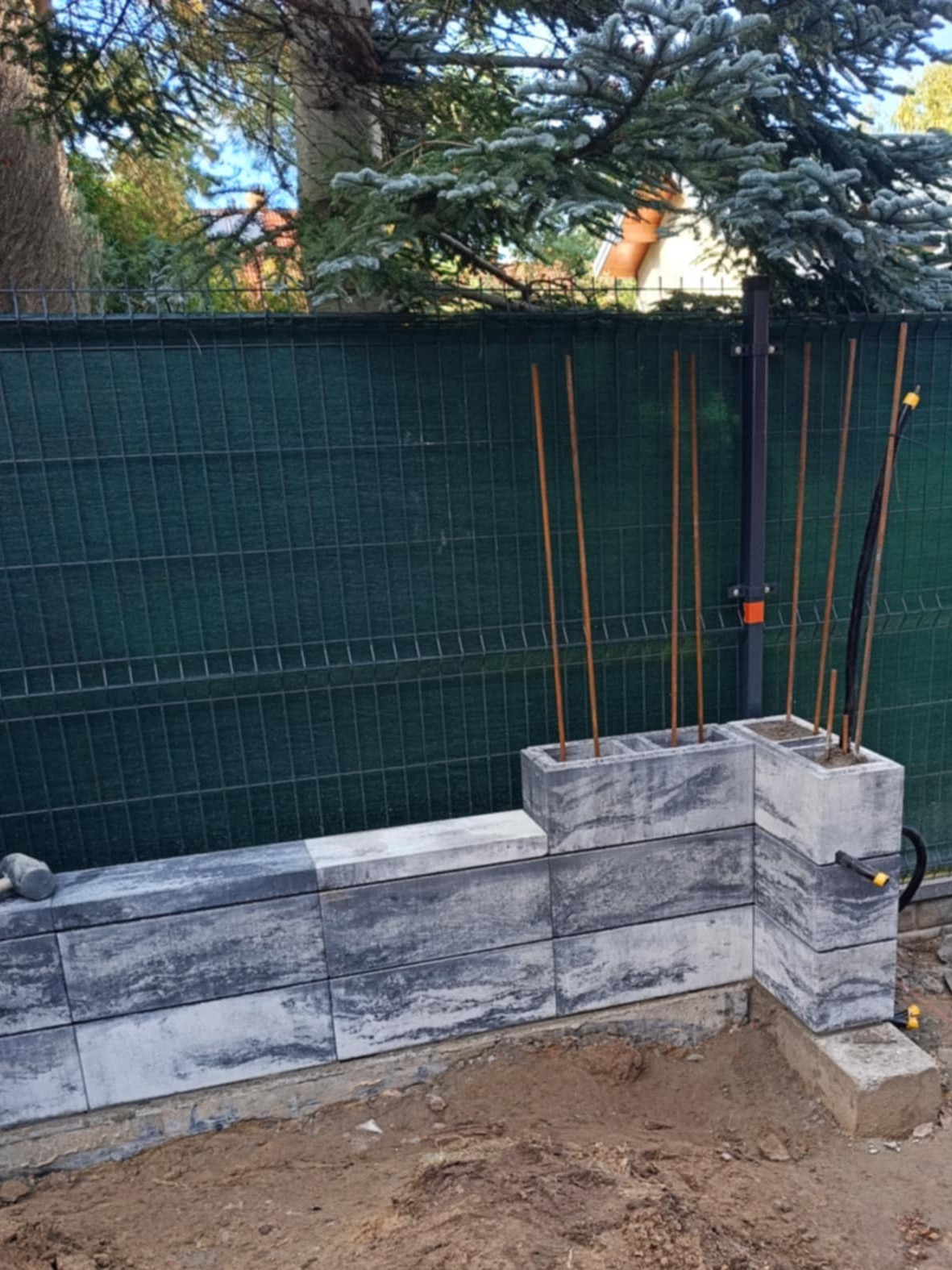 Wznoszenie murku z betonowych bloczków w kolorze szaro-białym, widoczne pręty zbrojeniowe wystające z pustaków, w tle zielona siatka ogrodzeniowa i drzewa.