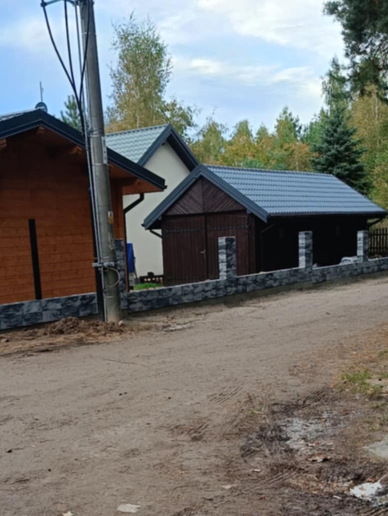 Nowoczesne ogrodzenie z kamiennych bloczków oddzielające posesję z drewnianym garażem i domem od piaszczystej drogi.
