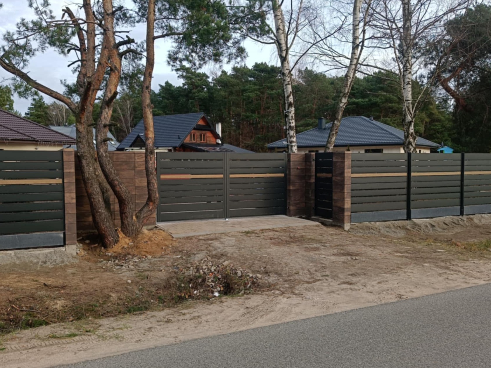 Nowoczesne ogrodzenie panelowe z bramą przesuwną i furtką, z elementami imitującymi drewno, osadzone na betonowej podmurówce i słupkach z cegły klinkierowej, z drzewami wkomponowanymi w linię...