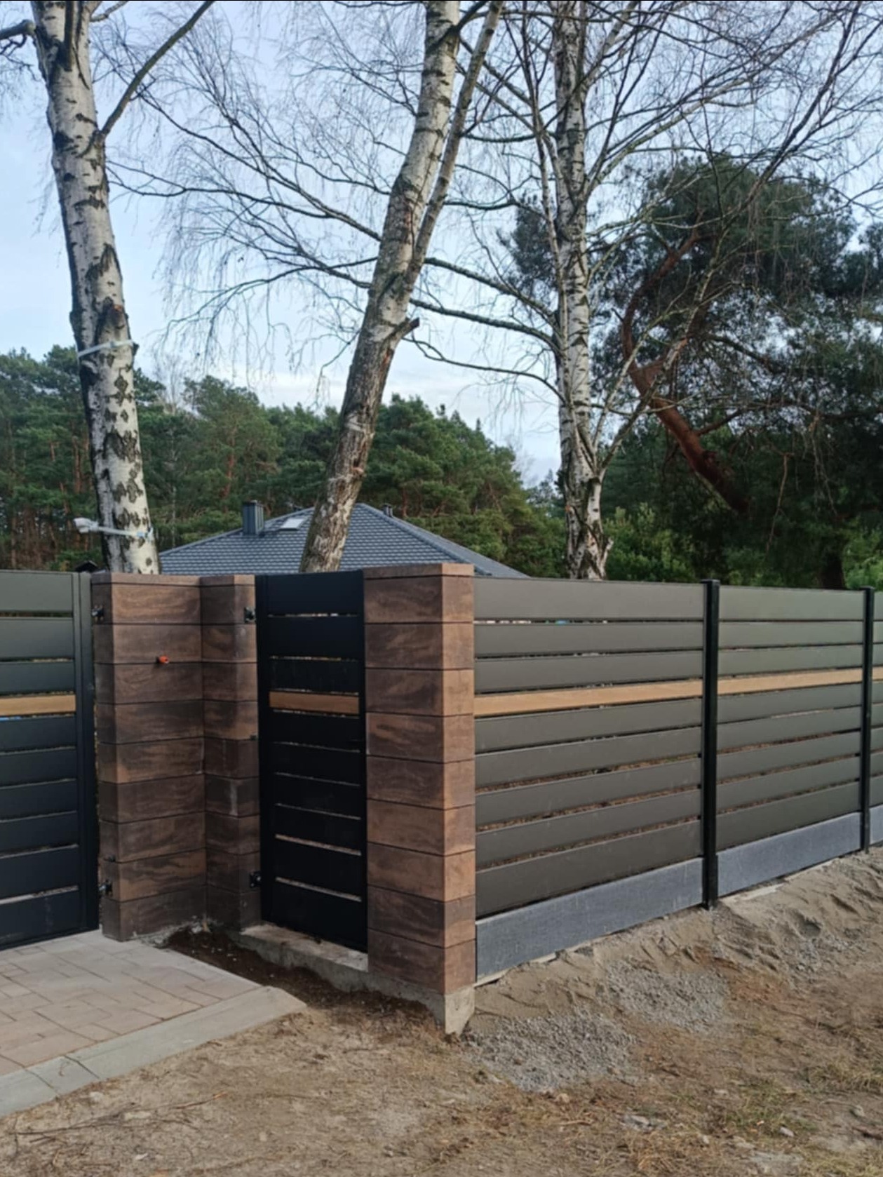 Nowoczesne ogrodzenie z poziomych paneli w kolorze grafitowym z elementami dekoracyjnymi w kolorze drewna, osadzone na słupkach wykończonych imitacją cegły, widok z boku na tle drzew i domu.