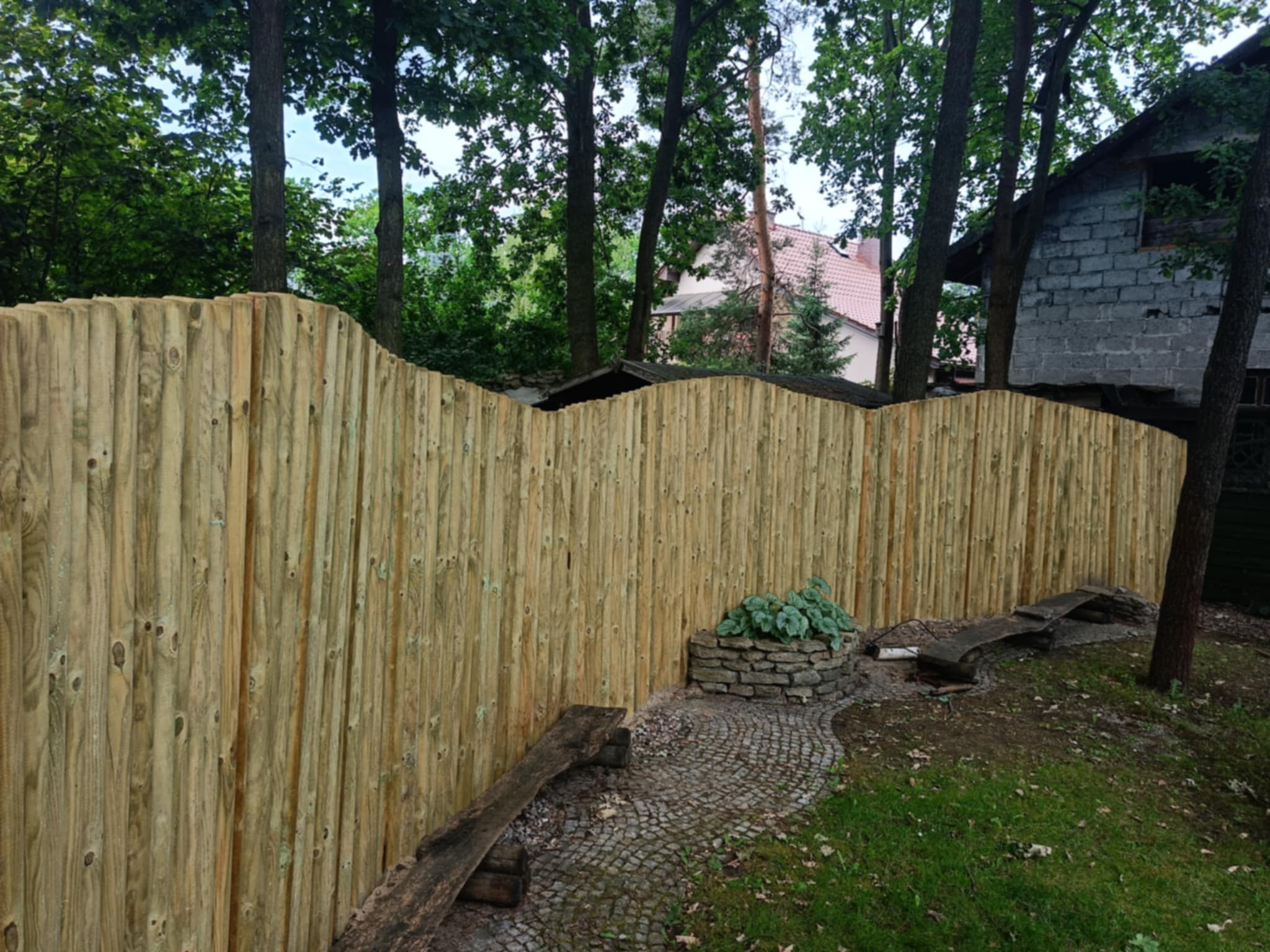 Nowe ogrodzenie z pionowych desek z zaokrąglonym wykończeniem, widok z zewnątrz, z fragmentem ścieżki z kostki brukowej i kamienną rabatką z roślinami.