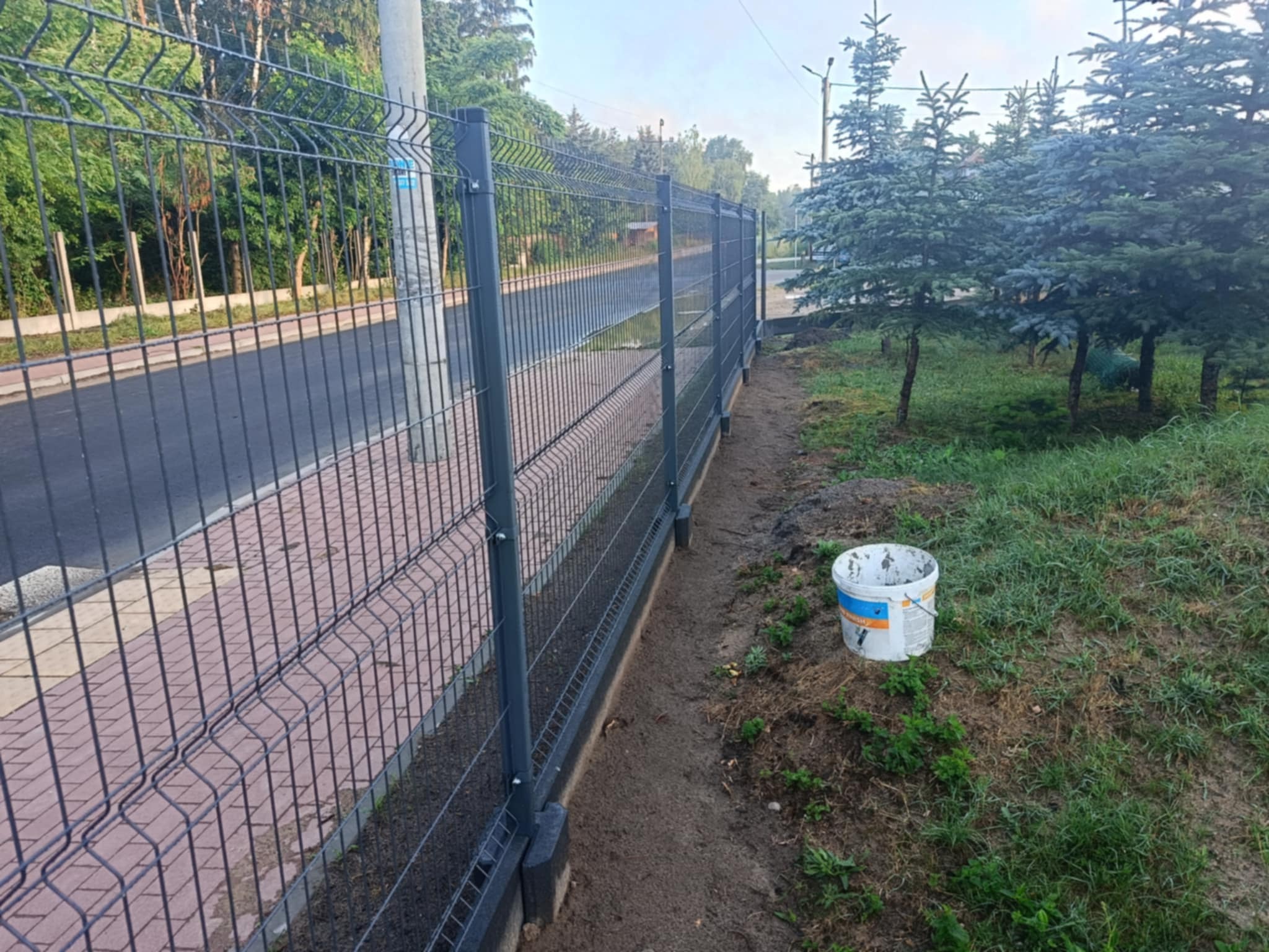 Zamontowane, grafitowe ogrodzenie panelowe przy chodniku z kostki brukowej, z widocznym wiadrem po farbie obok słupka.