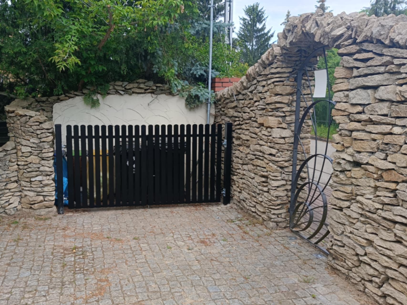 Czarna, przesuwna brama panelowa z pionowych sztachet, wkomponowana w kamienny mur z łukowym przejściem, na podjeździe z kostki brukowej.
