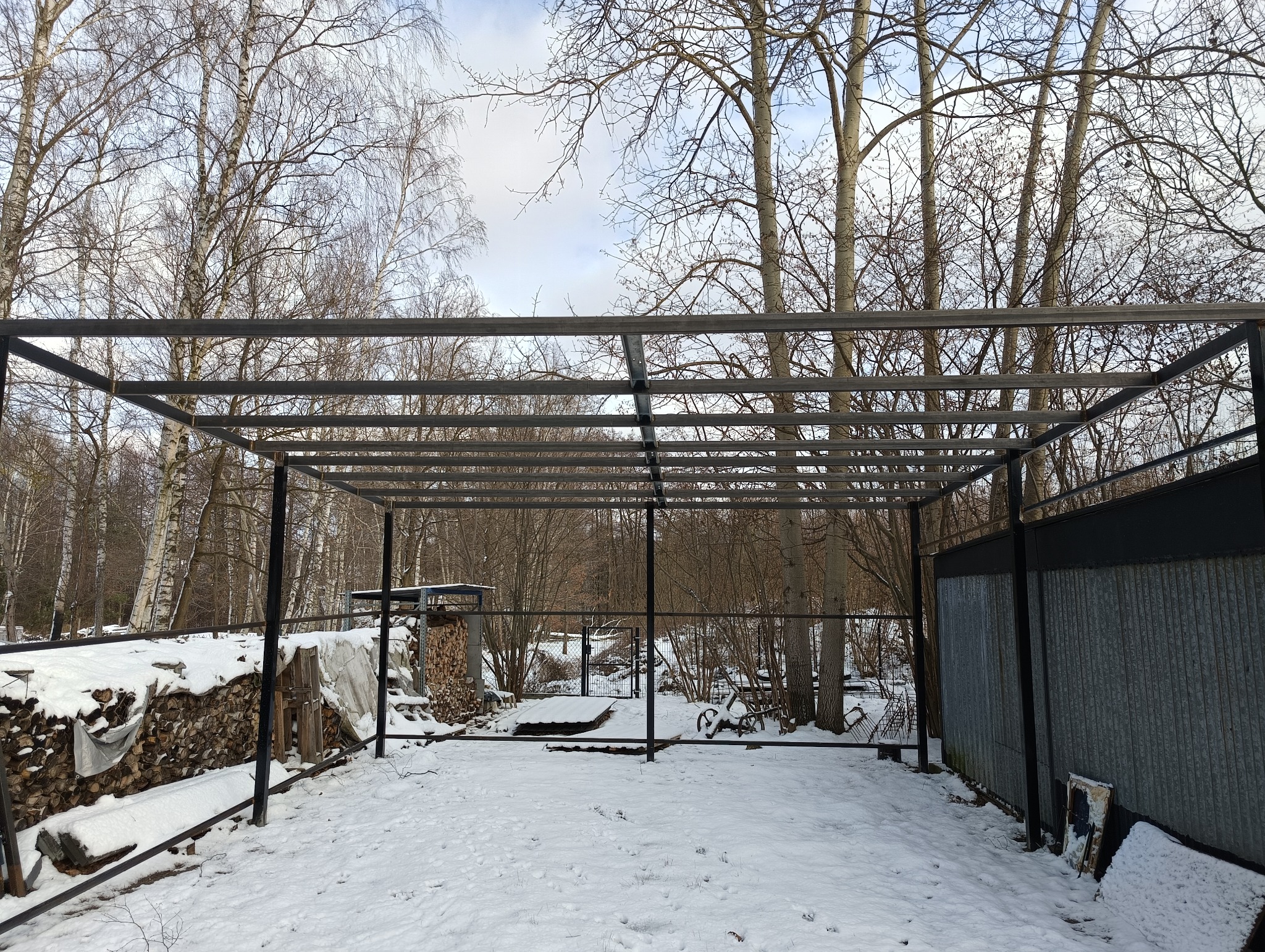 Szkielet metalowej konstrukcji w zimowym krajobrazie, z widocznym stosem drewna i częścią blaszanej ściany w tle.