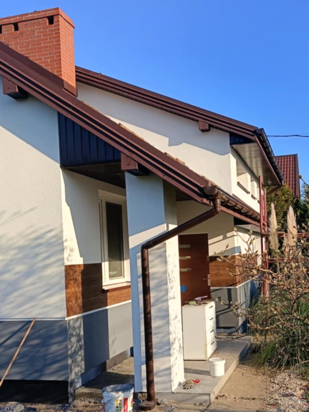 Elewacja domu jednorodzinnego w trakcie odnawiania: widoczna świeżo pomalowana ściana, brązowe rynny, fragment okna, drzwi wejściowe i rozstawione narzędzia malarskie.
