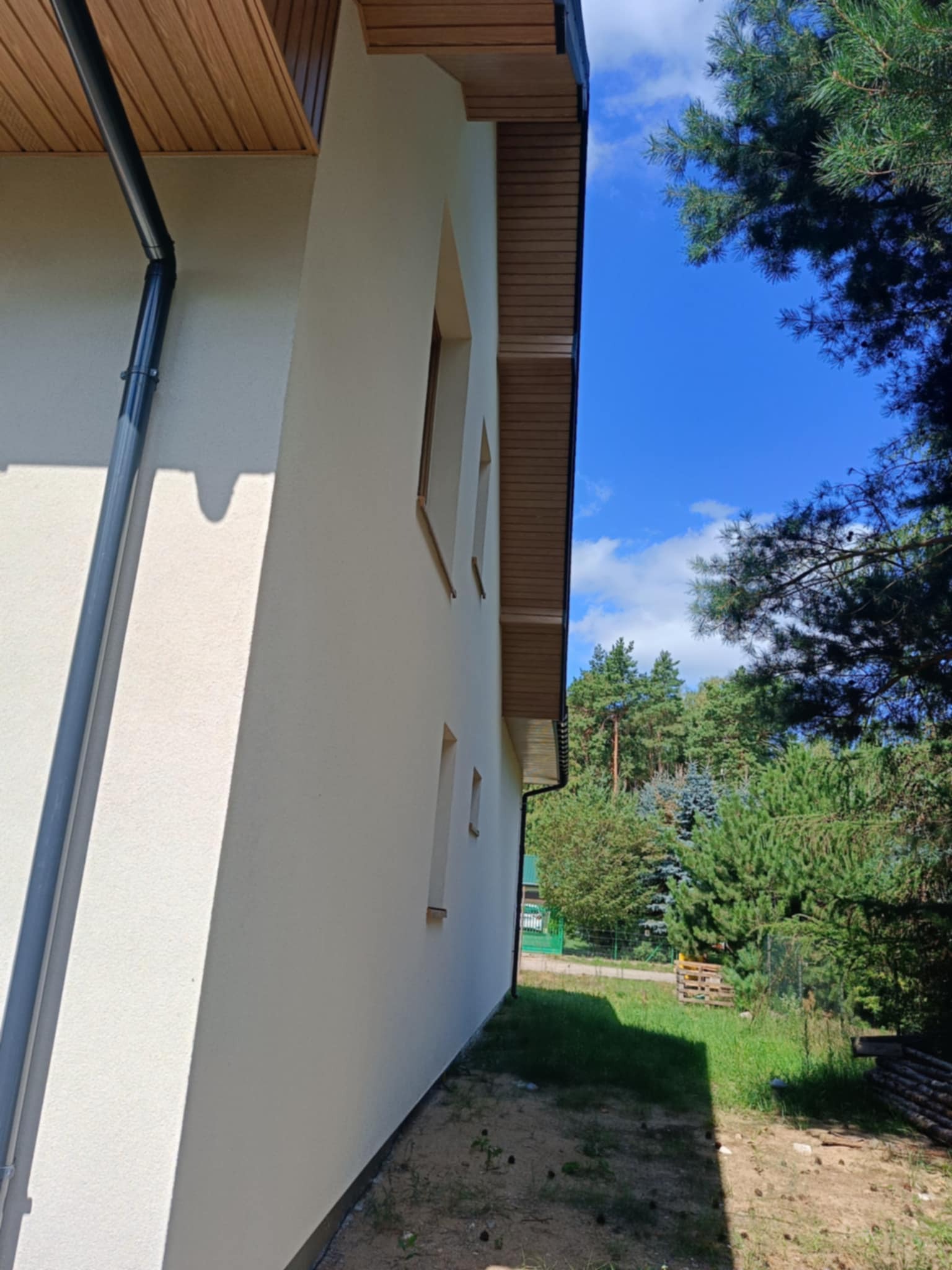 Jasna elewacja domu jednorodzinnego z drewnianą podbitką dachową i szarą rynną, widok z dołu na tle błękitnego nieba i zieleni drzew.