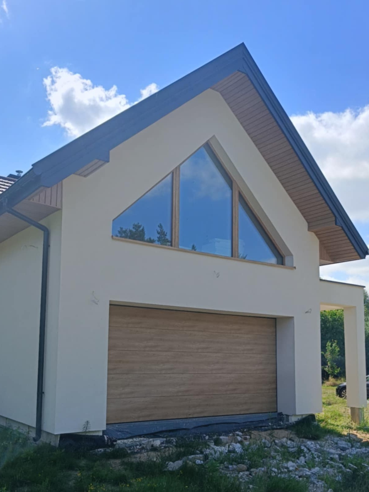 Nowoczesny dom z jasną elewacją, drewnianą bramą garażową segmentową i trójkątnym oknem na poddaszu odbijającym błękitne niebo.