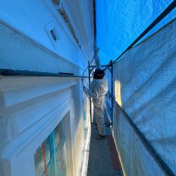 ZAKŁAD WIRTUALNEJ RZECZYWISTOŚCI-The Property of Virtual Universe Piotr Kuśmierski - Pracownik w kombinezonie ochronnym maluje elewację budynku w Warszawie za pomocą pistoletu natryskowego, stojąc na rusztowaniu zabezpieczonym siatką i plandeką.