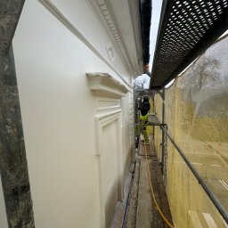 ZAKŁAD WIRTUALNEJ RZECZYWISTOŚCI-The Property of Virtual Universe Piotr Kuśmierski - Pracownik w kombinezonie ochronnym maluje elewację domu z dekoracyjnymi elementami architektonicznymi, stojąc na rusztowaniu zabezpieczonym siatką.