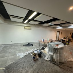 ZAKŁAD WIRTUALNEJ RZECZYWISTOŚCI-The Property of Virtual Universe Piotr Kuśmierski - Przestronne biuro w trakcie malowania, podłoga zabezpieczona folią, widoczne wiadra z farbą i narzędzia malarskie, sufit z panelami akustycznymi i oświetleniem LED.