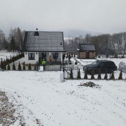 ZAKŁAD WIRTUALNEJ RZECZYWISTOŚCI-The Property of Virtual Universe Piotr Kuśmierski - Zimowy krajobraz z nowoczesnym domem jednorodzinnym z czarną dachówką, ogrodzeniem z tujami i drewnianą drewutnią w tle. Widoczny samochód i częściowo zasypany śniegiem teren.