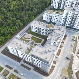 ZAKŁAD WIRTUALNEJ RZECZYWISTOŚCI-The Property of Virtual Universe Piotr Kuśmierski - Nowoczesne osiedle z lotu ptaka: białe budynki wielorodzinne, zielone dachy, plac zabaw i uporządkowana przestrzeń wokół. Widok z góry na architekturę i zagospodarowanie terenu.