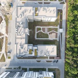 ArchUp - Kompleks budynków mieszkalnych z zielonym dziedzińcem, placem zabaw i parkingiem, widziany z lotu ptaka. Obok las. Częściowo widoczny plac budowy.