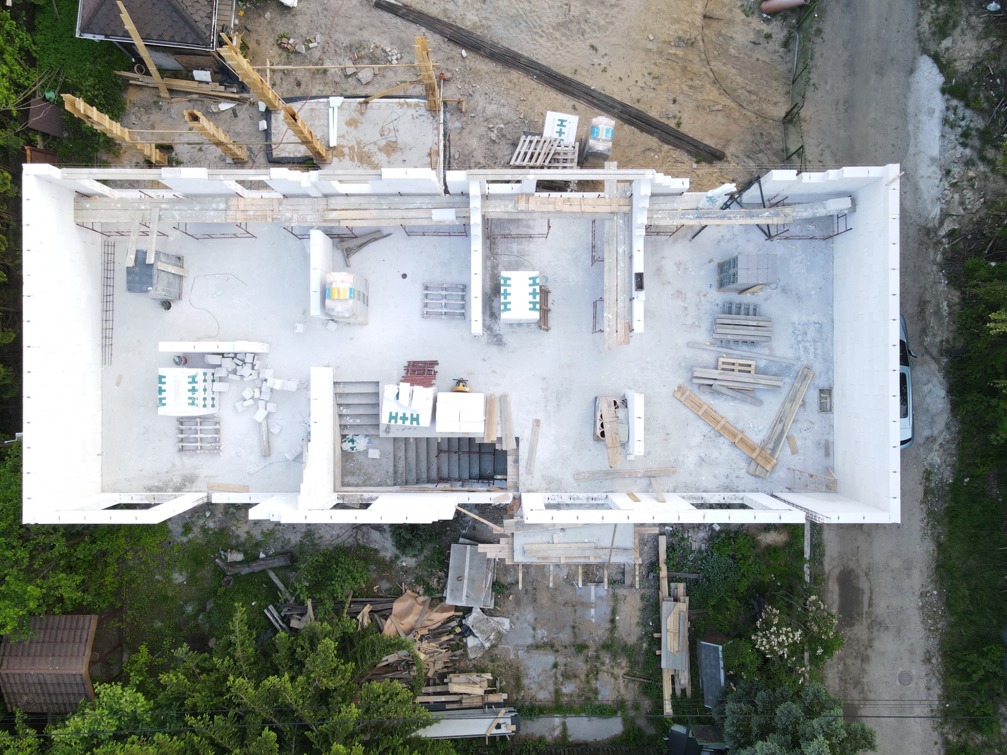 Budowa domu jednorodzinnego z pustaków H+H, widok z drona. Wnętrze w stanie surowym otwartym, widoczne schody i elementy konstrukcyjne dachu. Otoczenie: zieleń, piasek, materiały budowlane.
