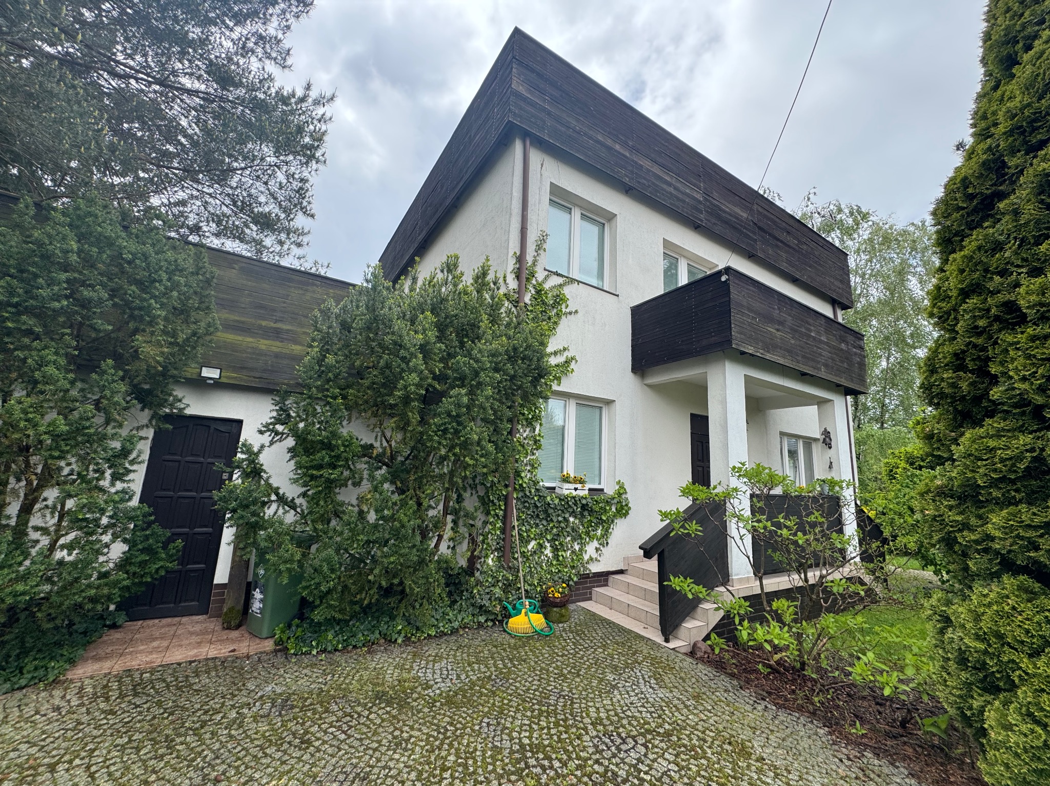 Dwupiętrowy dom z balkonem, białą elewacją i ciemnym drewnianym wykończeniem dachu oraz balkonu. Na podjeździe leży żółty wąż ogrodowy i grabie.