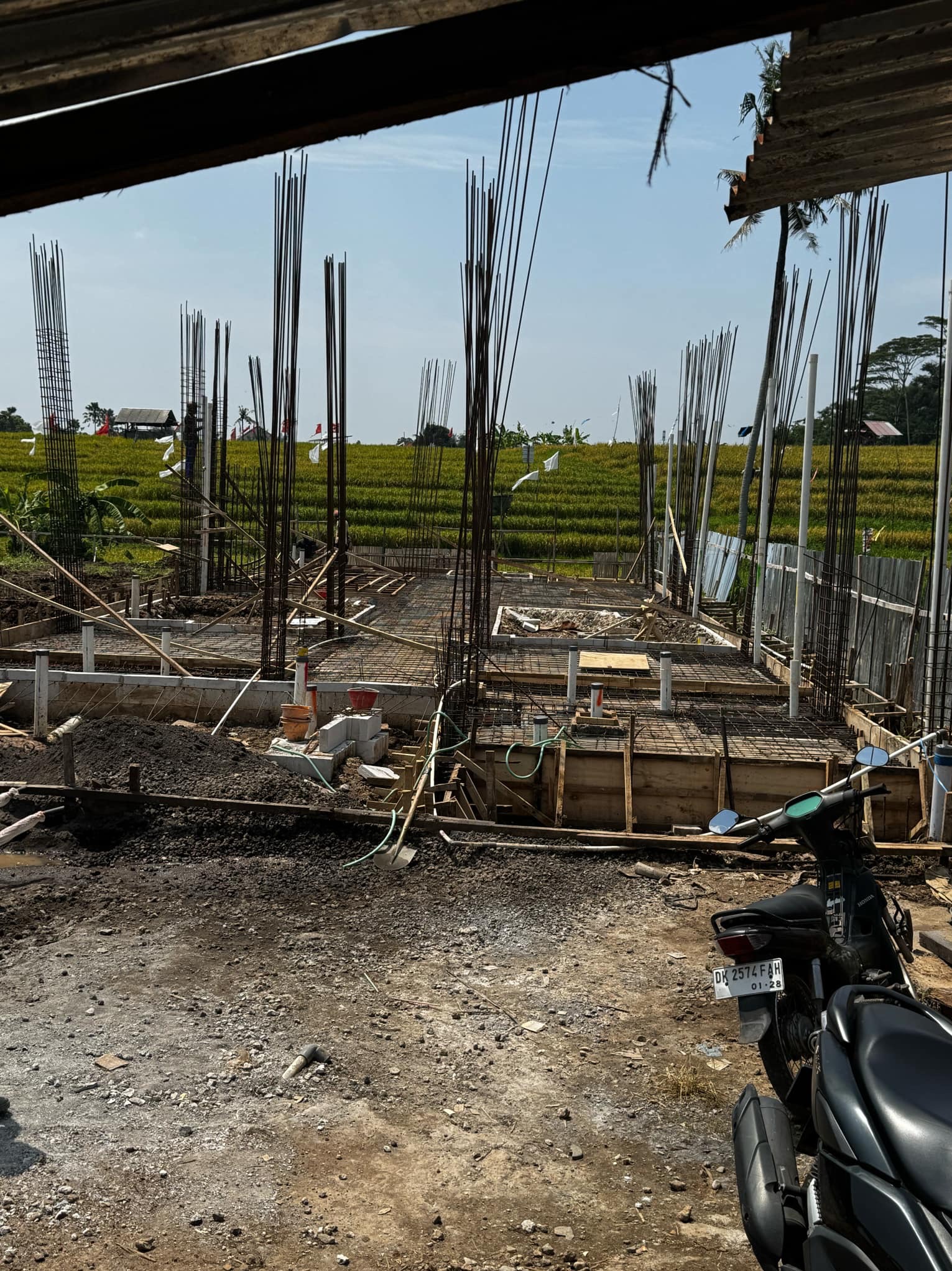 Budowa domu w trakcie: fundamenty zbrojone stalą na tle zielonego pola ryżowego. Widoczne szalunki, pionowe pręty zbrojeniowe i motocykl w rogu kadru.