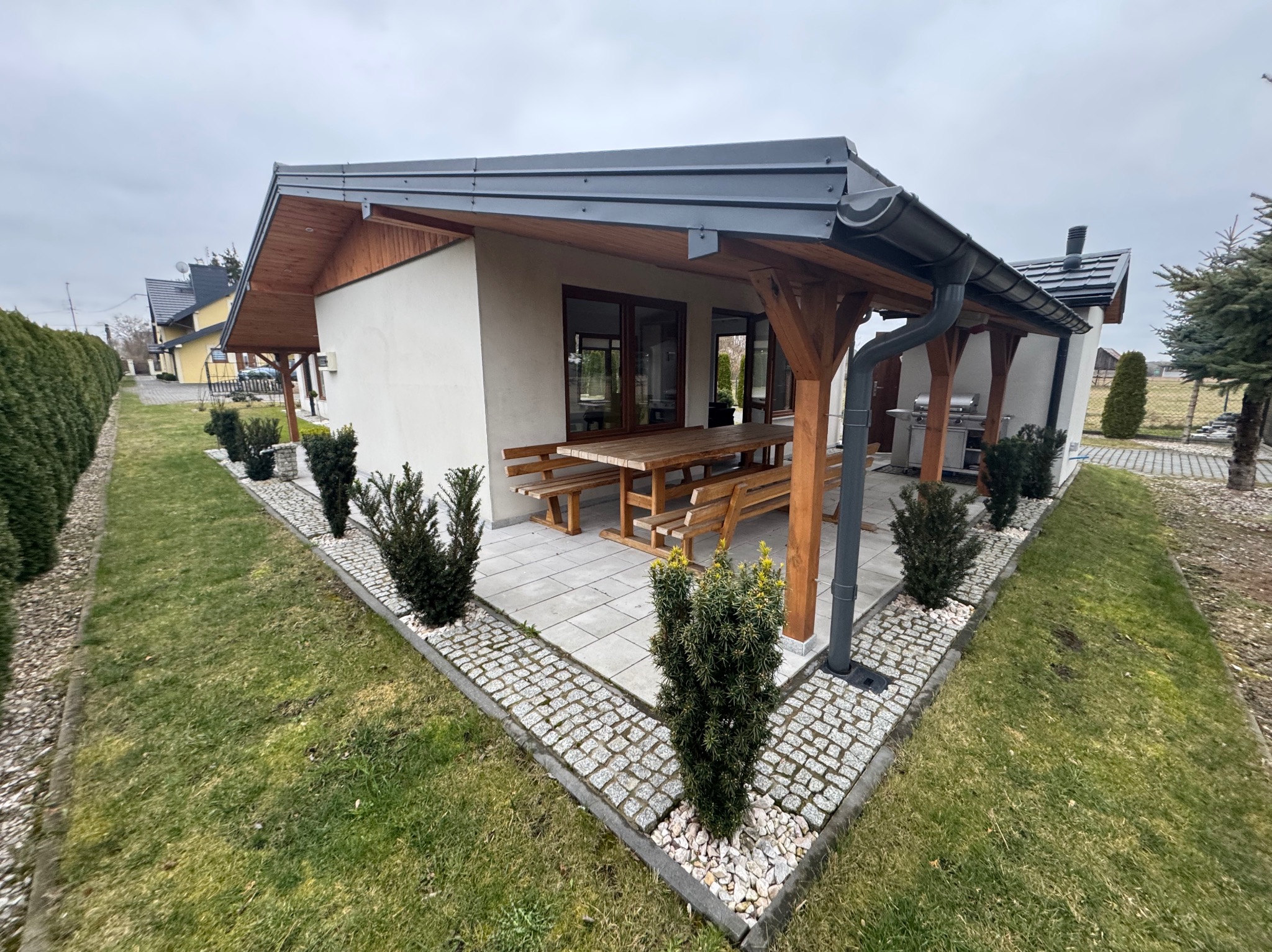 Nowoczesny dom z tarasem i drewnianą pergolą, otoczony zielenią. Widoczny stół z ławkami i grill. Całość utrzymana w stonowanej kolorystyce.