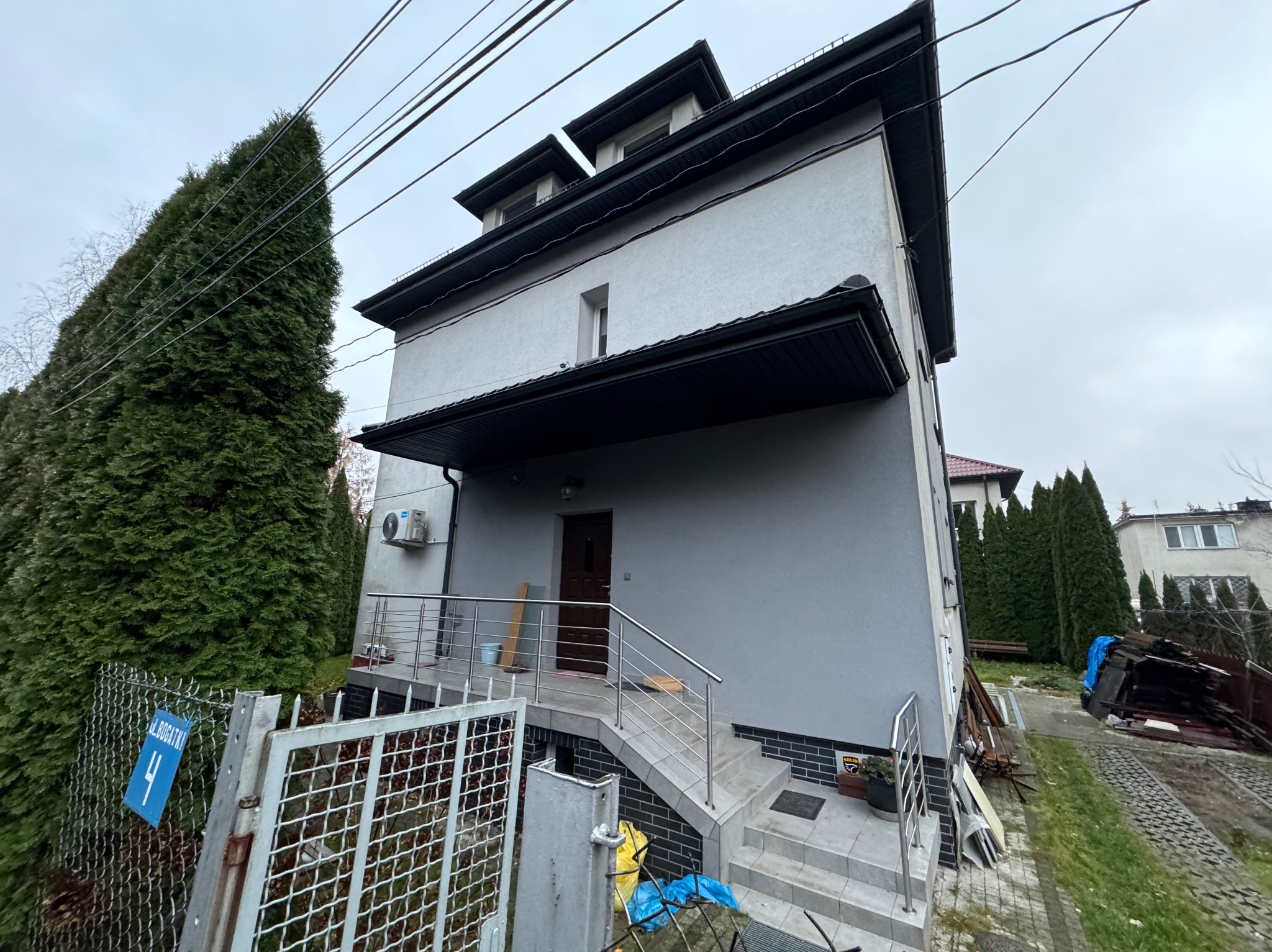 Szary, dwupiętrowy dom z poddaszem użytkowym, widziany z dołu, z metalową balustradą i schodami wejściowymi. Na ogrodzeniu niebieski numer domu.