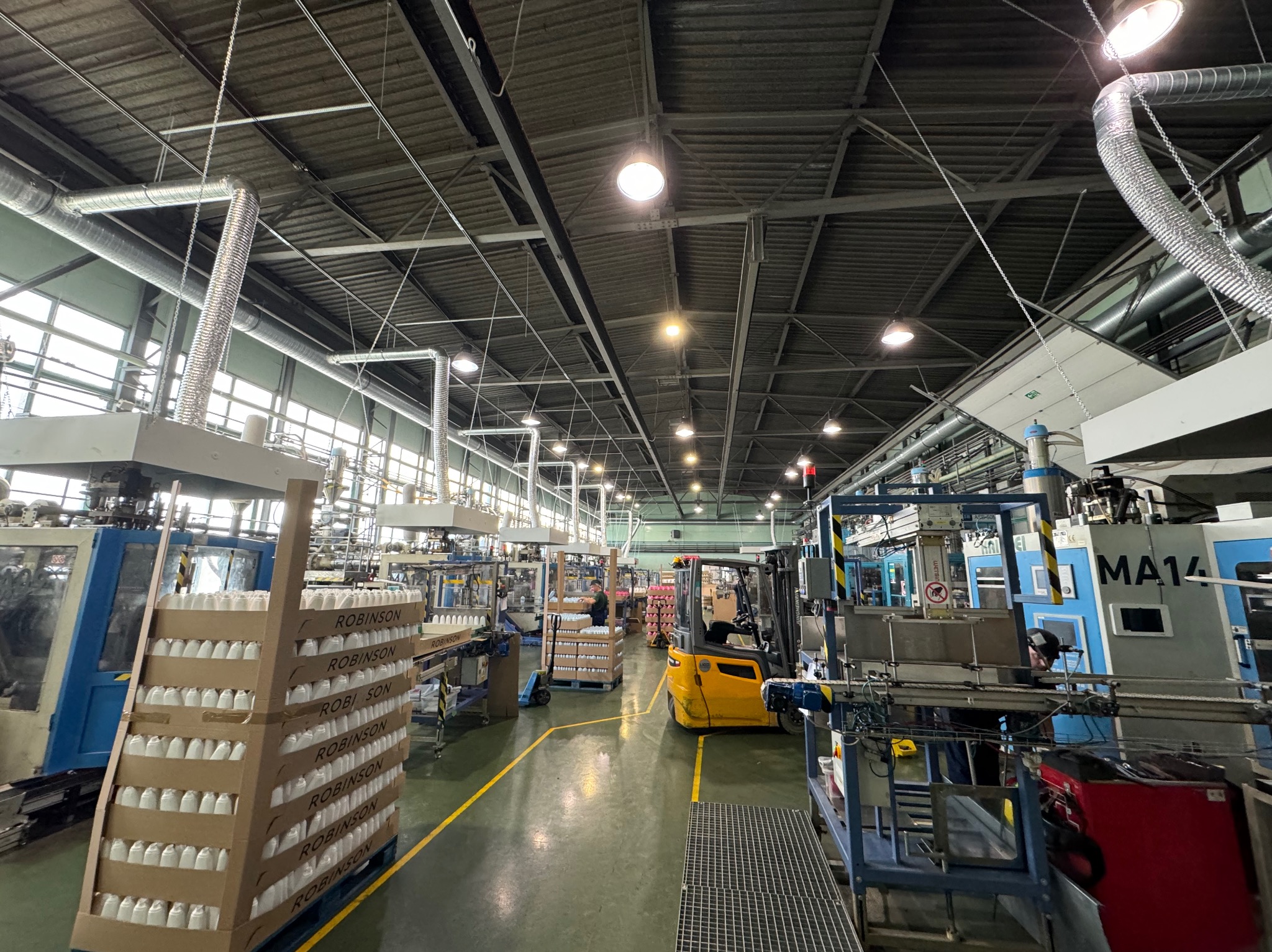 Wnętrze hali produkcyjnej z liniami technologicznymi, wózkiem widłowym i pracownikami. Widoczna instalacja wentylacyjna pod sufitem. Produkcja opakowań.
