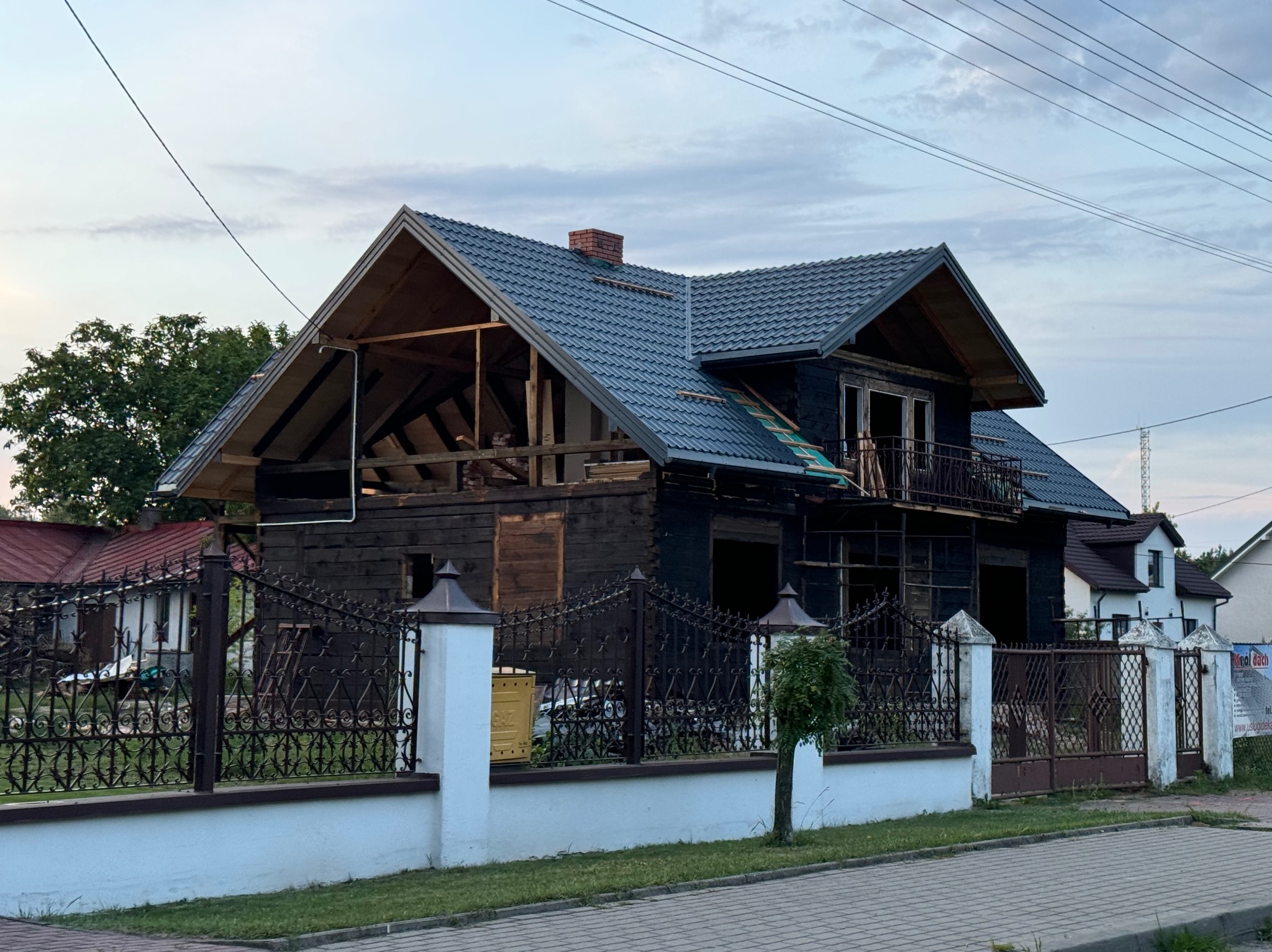Dom w trakcie przebudowy z widoczną konstrukcją dachu i nowym pokryciem. Czarny kolor elewacji kontrastuje z białą podmurówką ogrodzenia.