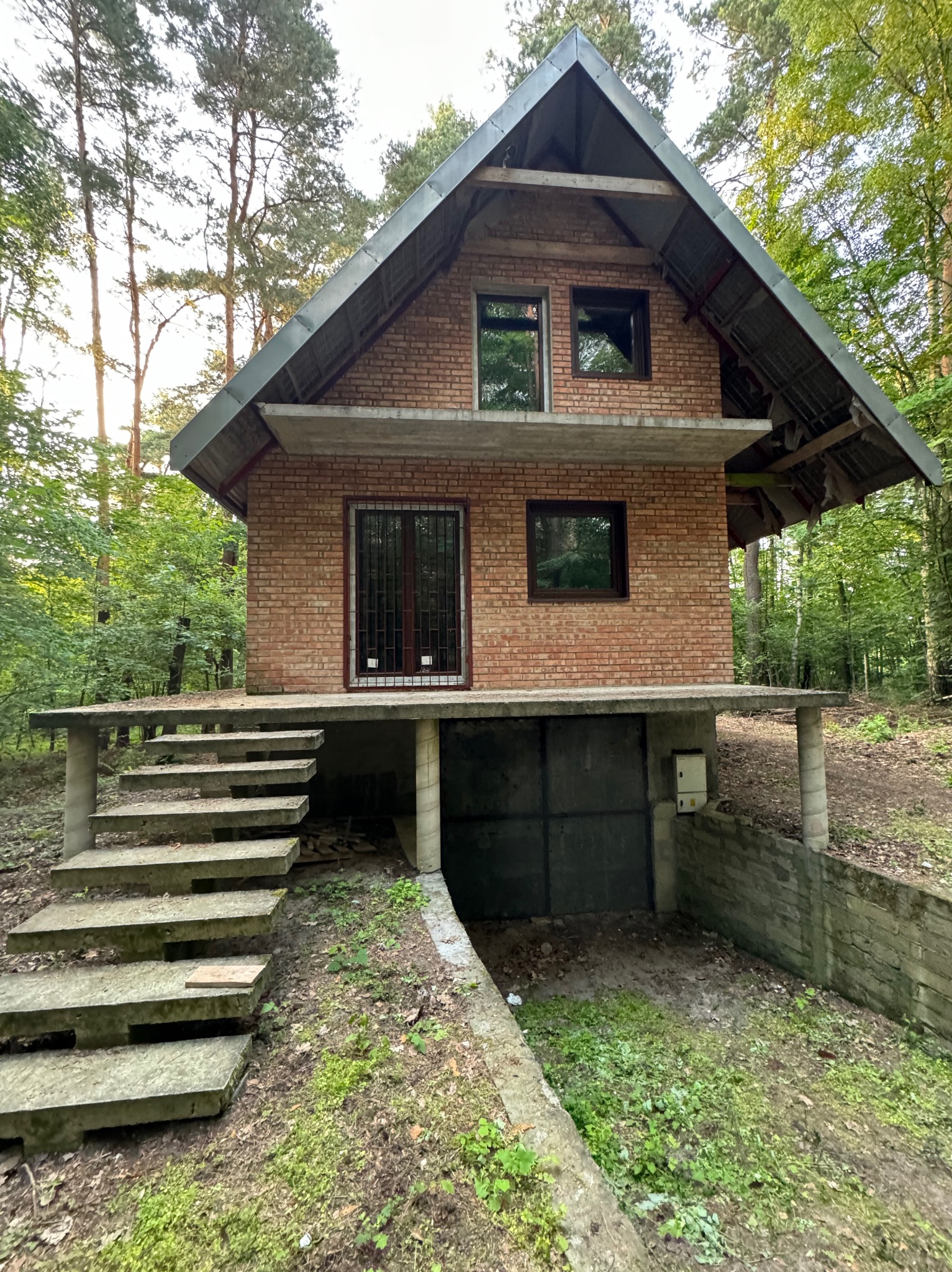 Niedokończony, dwukondygnacyjny dom z cegły w leśnej scenerii. Widoczne betonowe schody i podpiwniczenie. Surowy stan budynku, otoczenie leśne.