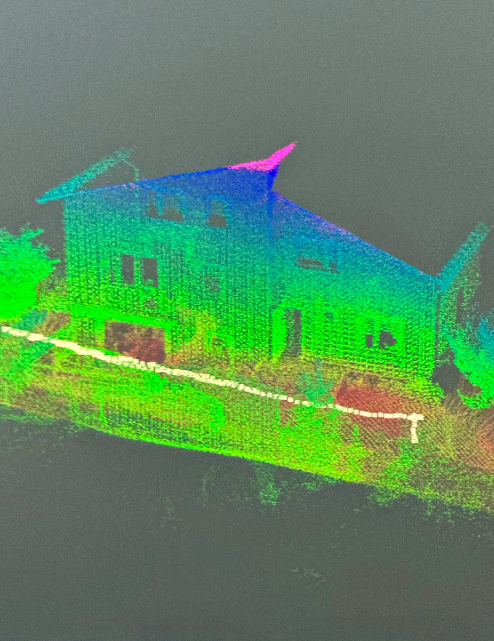 Skan 3D domu z kolorową wizualizacją punktów, ukazujący bryłę budynku, okna i otoczenie w odcieniach zieleni, niebieskiego i różu na ciemnym tle. Unikalny widok z perspektywy.