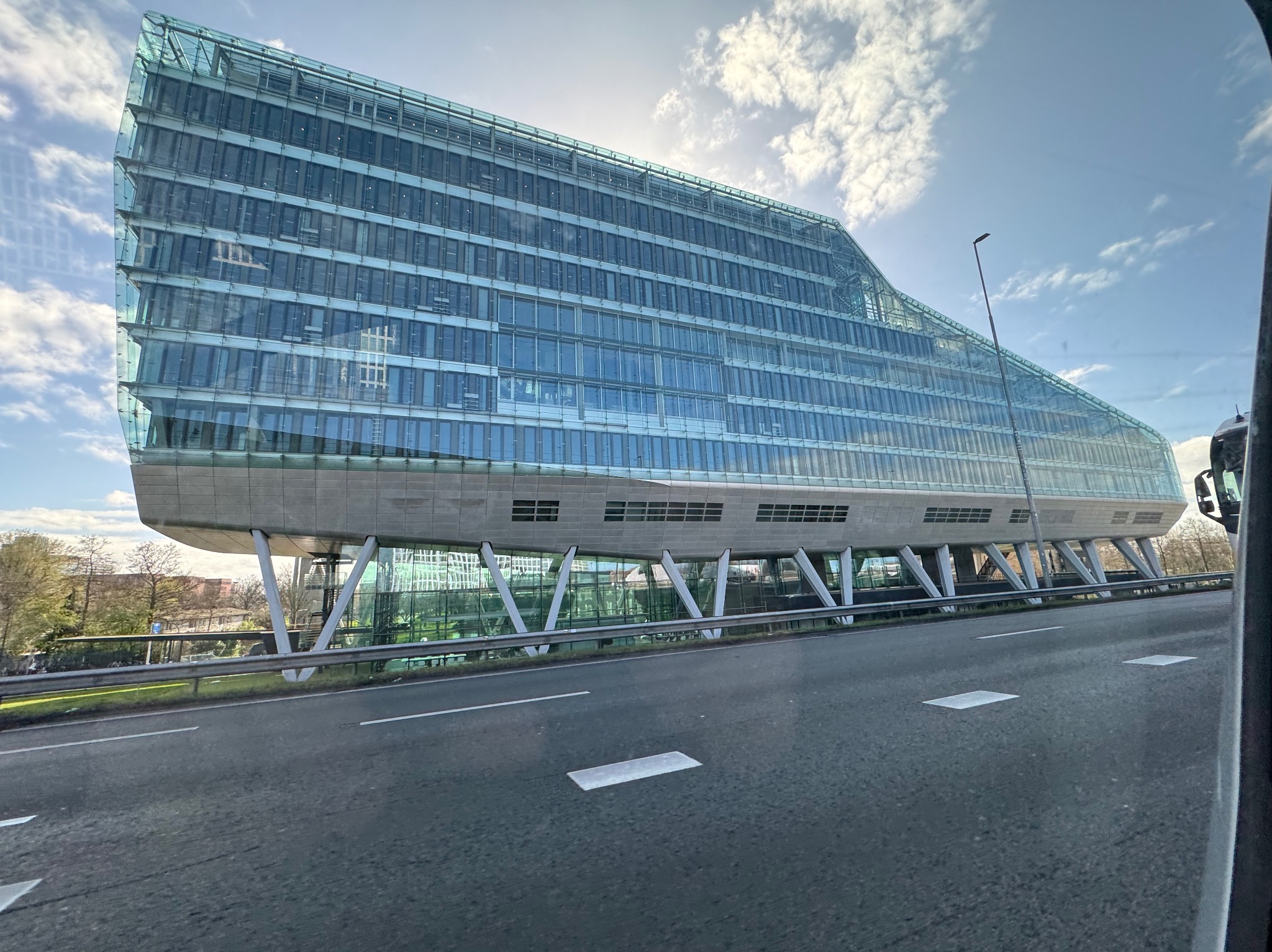 Nowoczesny biurowiec o szklanej fasadzie, wsparty na stalowych podporach, widziany z perspektywy jezdni. Błękitne niebo z chmurami w tle.
