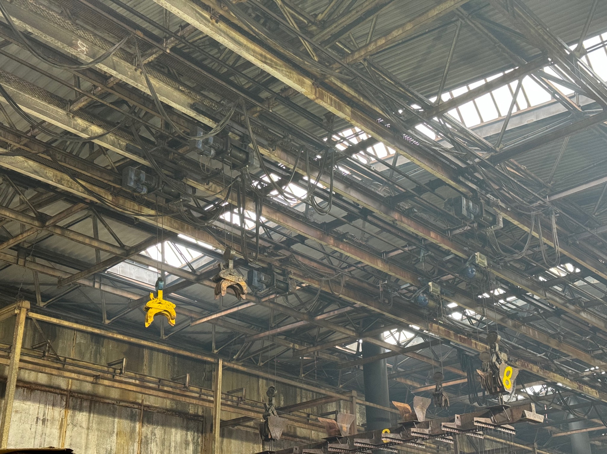 Wnętrze hali produkcyjnej z suwnicami i elementami konstrukcyjnymi pod dachem z świetlikami. Widoczne żółte haki suwnic i instalacje elektryczne.