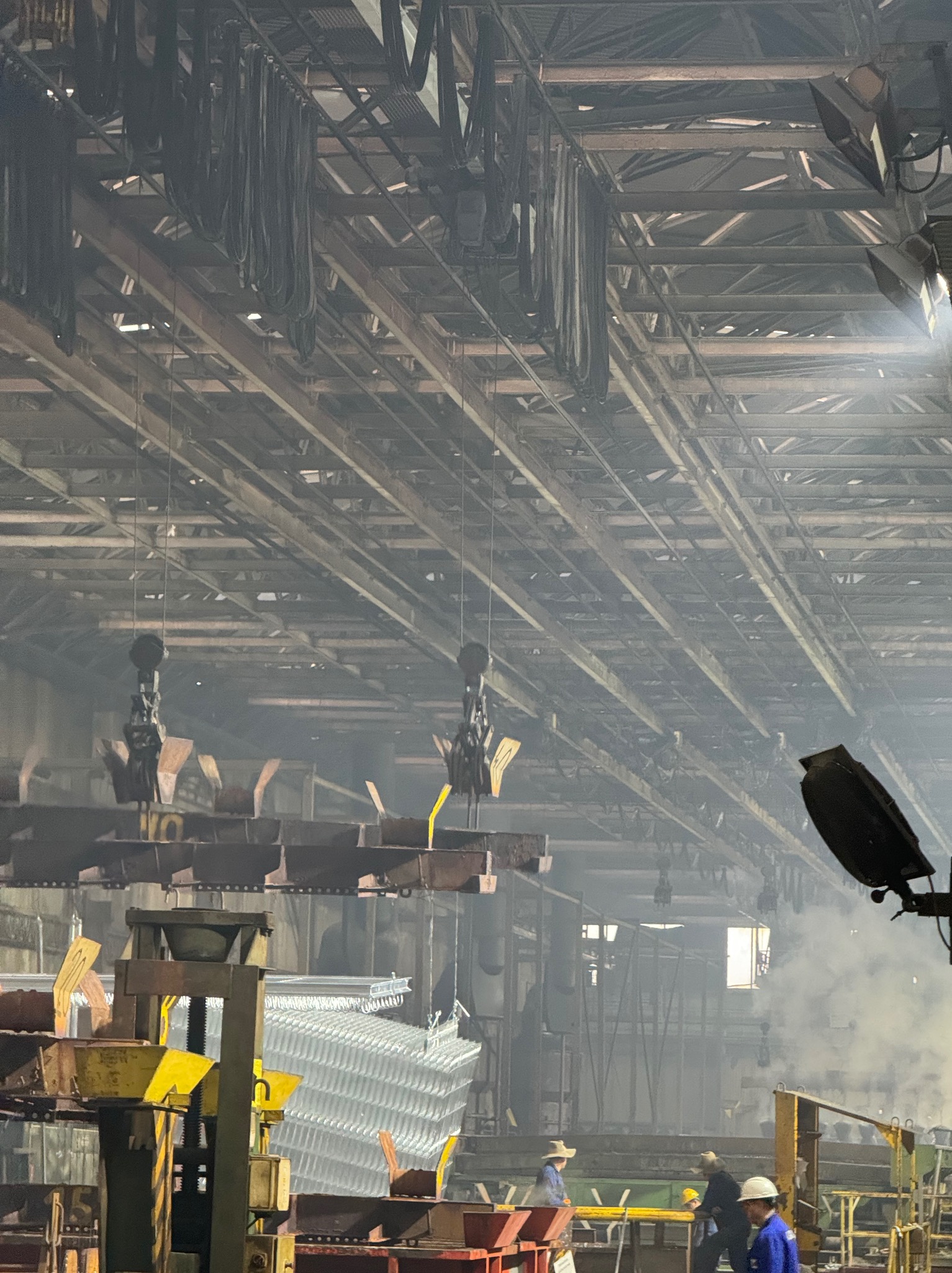 Wnętrze hali produkcyjnej z konstrukcją stalową, suwnicami i pracownikami w oddali. Widoczny proces obróbki metalu i unoszący się dym.