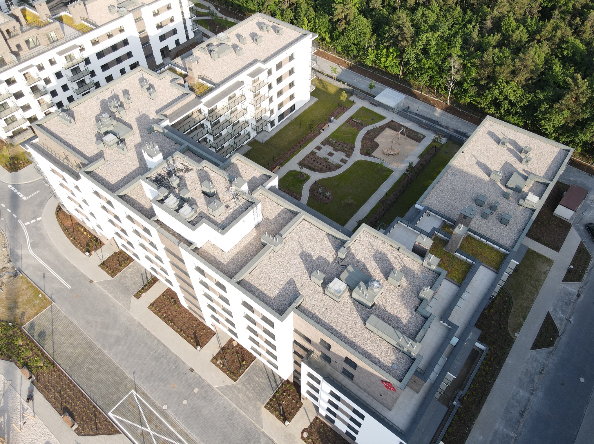 Nowoczesny kompleks budynków mieszkalnych z zielonym dziedzińcem i placem zabaw, widok z lotu ptaka, jasna kolorystyka elewacji, geometryczny układ dachów.