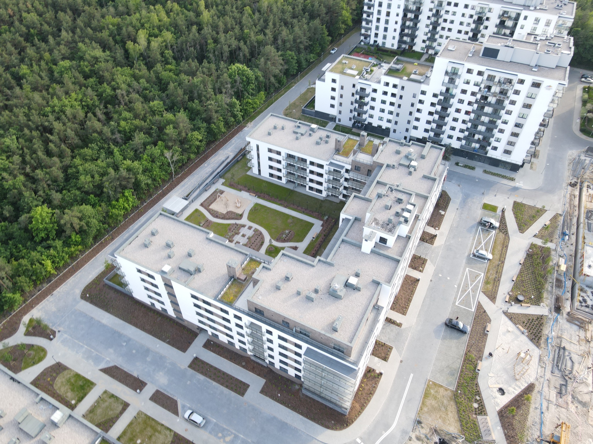 Nowoczesne osiedle z lotu ptaka: białe budynki wielorodzinne, zielone dachy, plac zabaw i uporządkowana przestrzeń wokół. Widok z góry na architekturę i zagospodarowanie terenu.