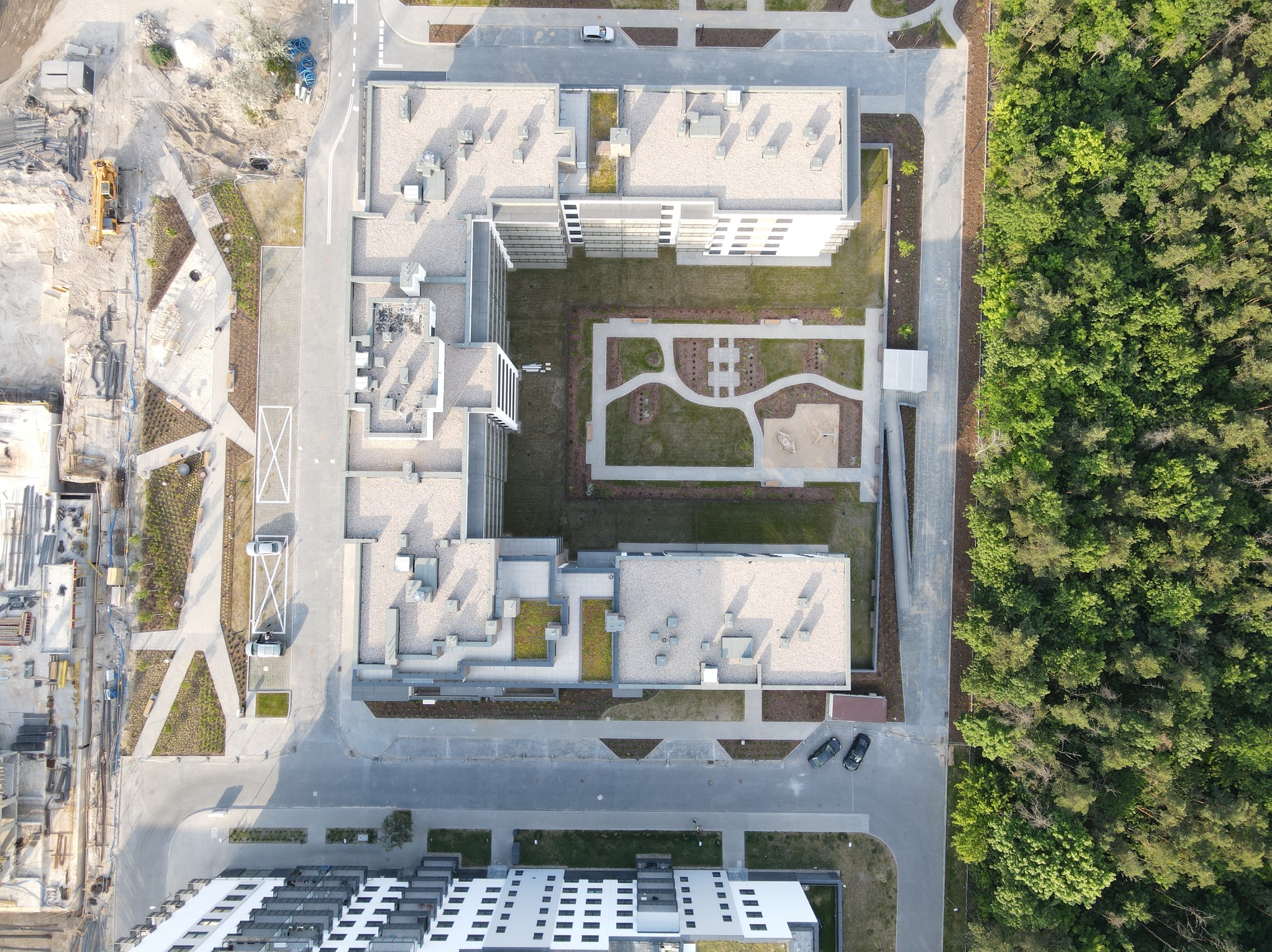 Kompleks budynków mieszkalnych z zielonym dziedzińcem, placem zabaw i parkingiem, widziany z lotu ptaka. Obok las. Częściowo widoczny plac budowy.