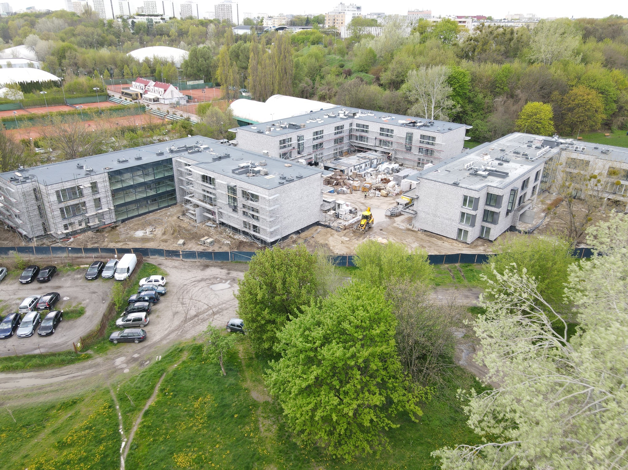 Wysoka perspektywa na kompleks budynków w trakcie budowy. Widoczne rusztowania, maszyny budowlane i parking z samochodami. W tle zieleń i zabudowa miejska.