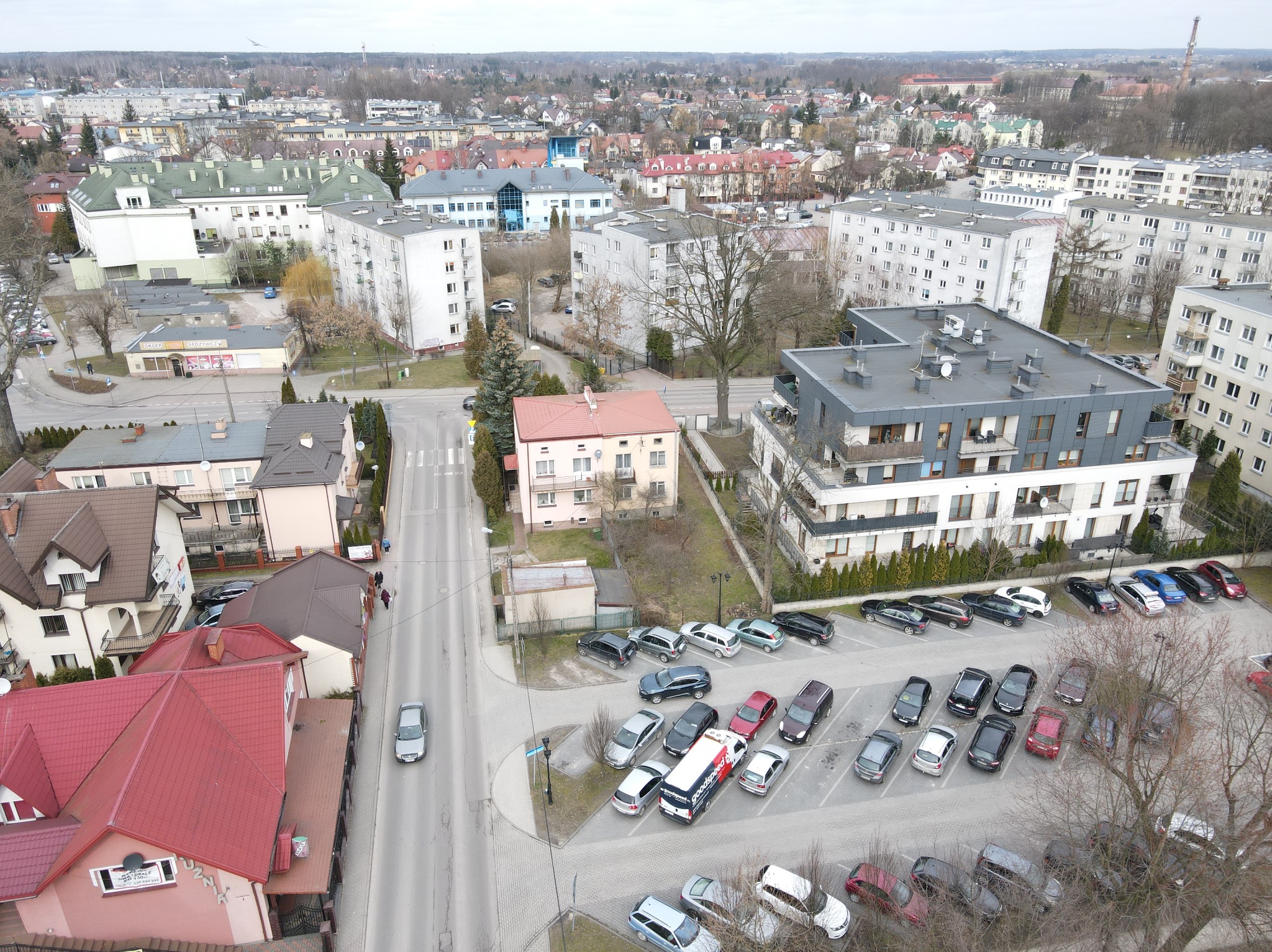 Widok z góry na miasto z blokami, domami i parkingiem pełnym aut. Ulica przecina zabudowę, a w oddali widoczne drzewa i budynki.
