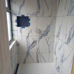 Paweł Grasco - Płytki imitujące marmur z niebieskimi żyłami na ścianach kabiny prysznicowej, z niebieską mozaiką heksagonalną w niszy i na podłodze. Widoczny biały brodzik i fragment okna.