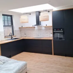 Nowoczesna kuchnia z czarnymi szafkami, drewnianym blatem i podłogą, oświetlona naturalnym światłem z okna dachowego, z dekoracyjnymi lampami wiszącymi i białymi płytkami na ścianie.