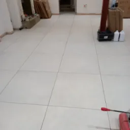 Ułożone jasnoszare płytki podłogowe z ciemnoszarą fugą, widoczny fragment przecinarki do płytek z czerwoną rączką, w tle niewykończone pomieszczenie z elementami dekoracyjnymi kominka i materiałami...