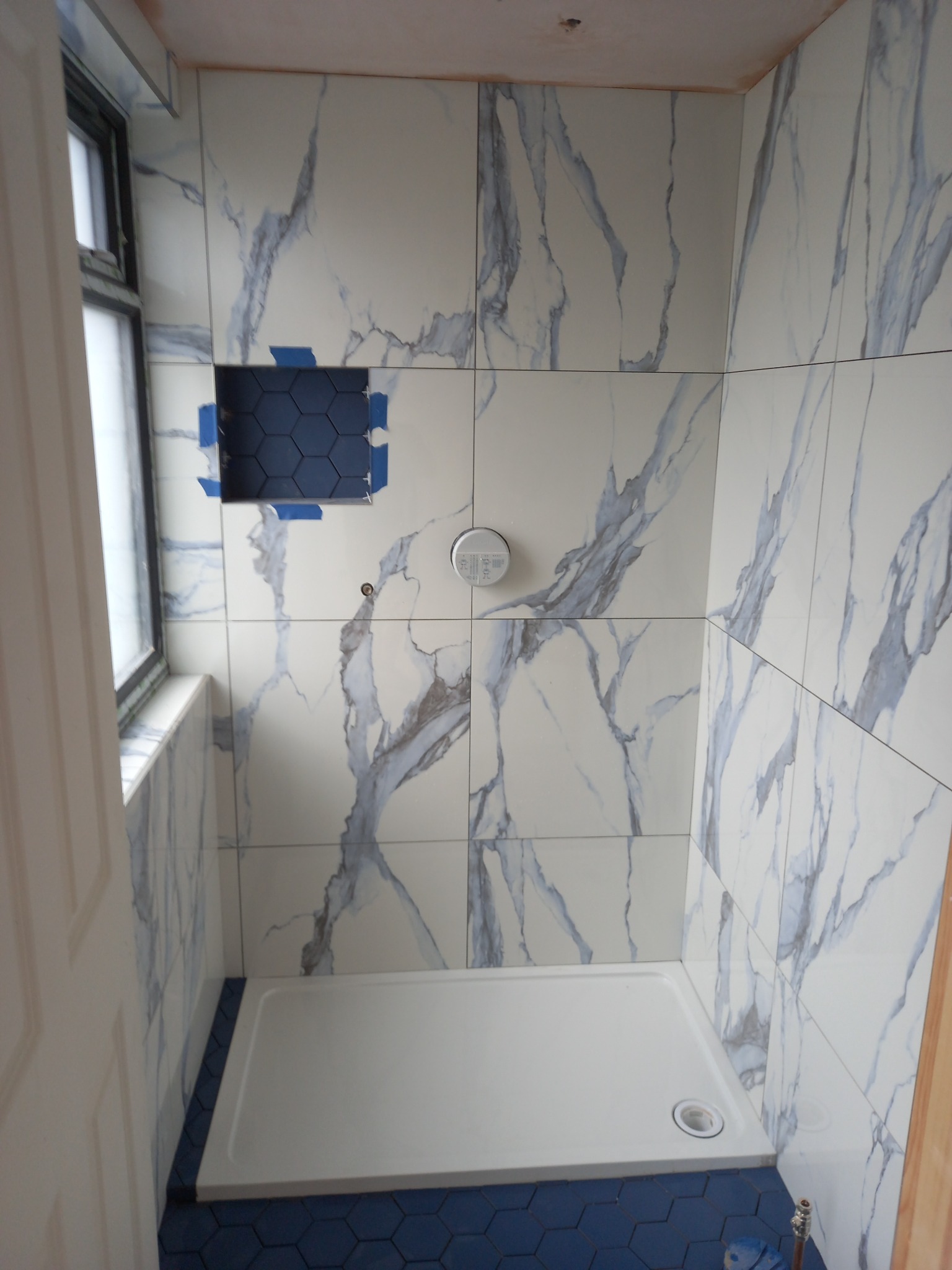 Płytki imitujące marmur z niebieskimi żyłami na ścianach kabiny prysznicowej, z niebieską mozaiką heksagonalną w niszy i na podłodze. Widoczny biały brodzik i fragment okna.