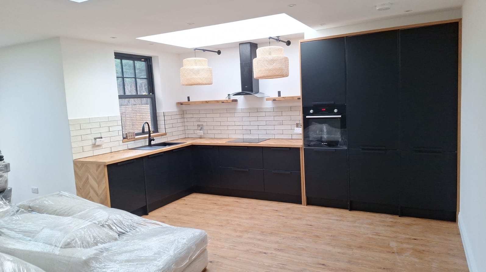 Nowoczesna kuchnia z czarnymi szafkami, drewnianym blatem i podłogą, oświetlona naturalnym światłem z okna dachowego, z dekoracyjnymi lampami wiszącymi i białymi płytkami na ścianie.