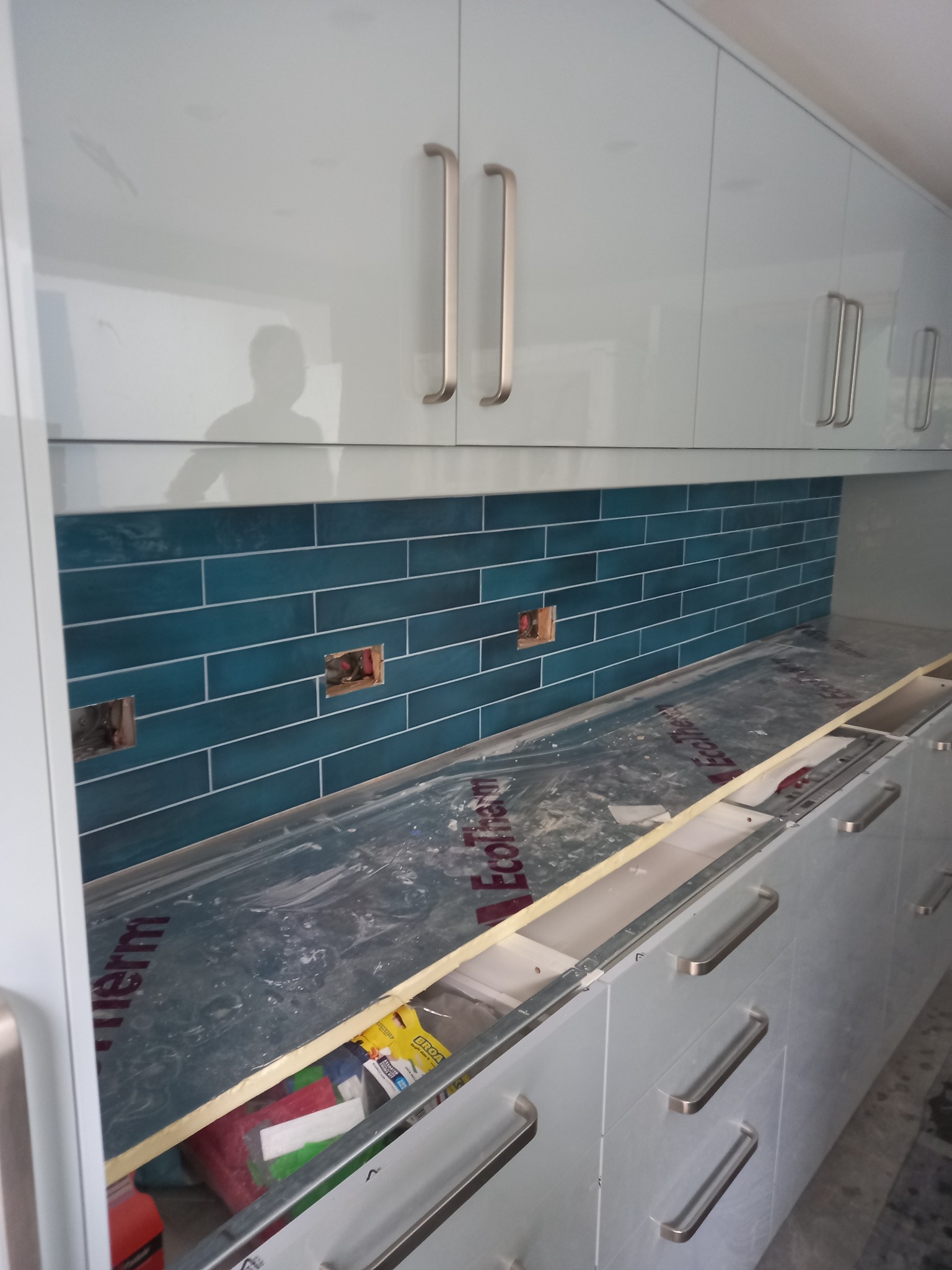 Montaż turkusowych płytek w kuchni, widoczne puszki elektryczne, meble kuchenne z połyskiem zabezpieczone folią.