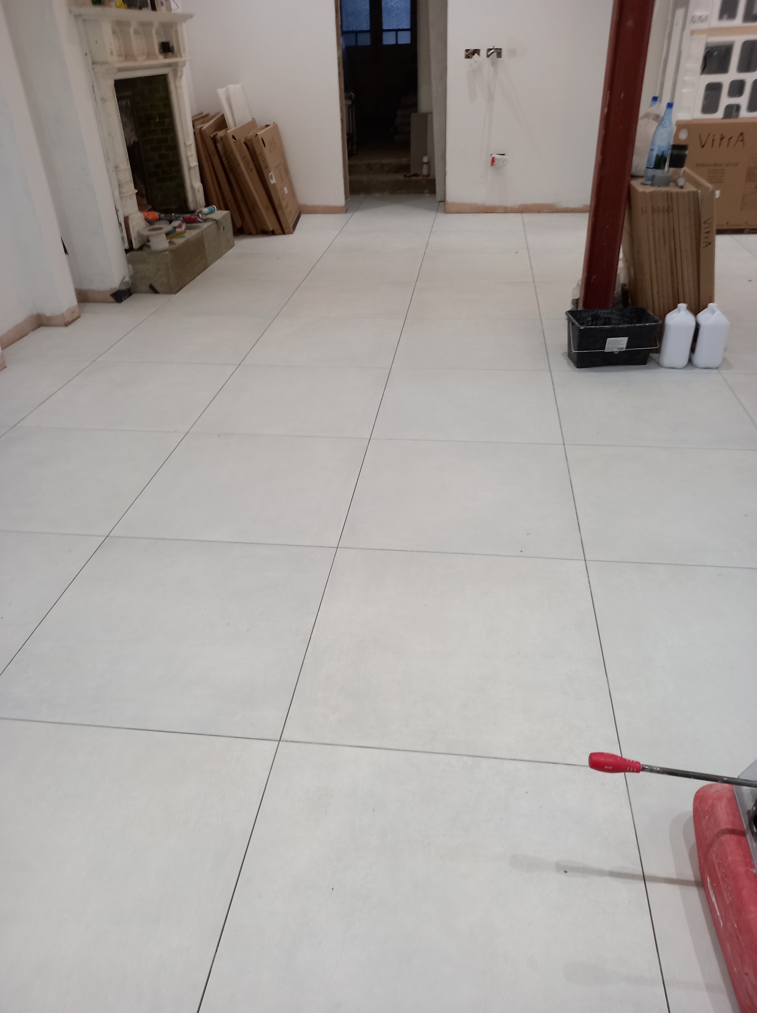 Ułożone jasnoszare płytki podłogowe z ciemnoszarą fugą, widoczny fragment przecinarki do płytek z czerwoną rączką, w tle niewykończone pomieszczenie z elementami dekoracyjnymi kominka i materiałami...