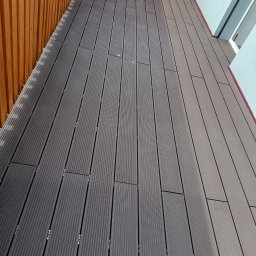 Dam-Bud Damian Przybylak - Balkon z szarych desek kompozytowych z widocznymi mocowaniami, obok drewniana ażurowa ścianka i biała ściana budynku z oknem.